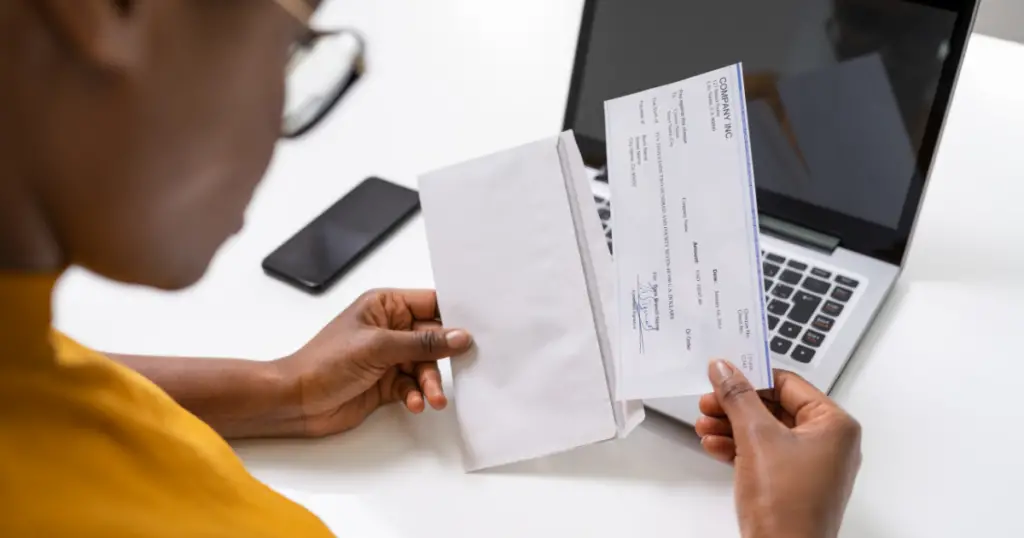 Person pulling a check out of an envelope with a laptop and cell phone in background