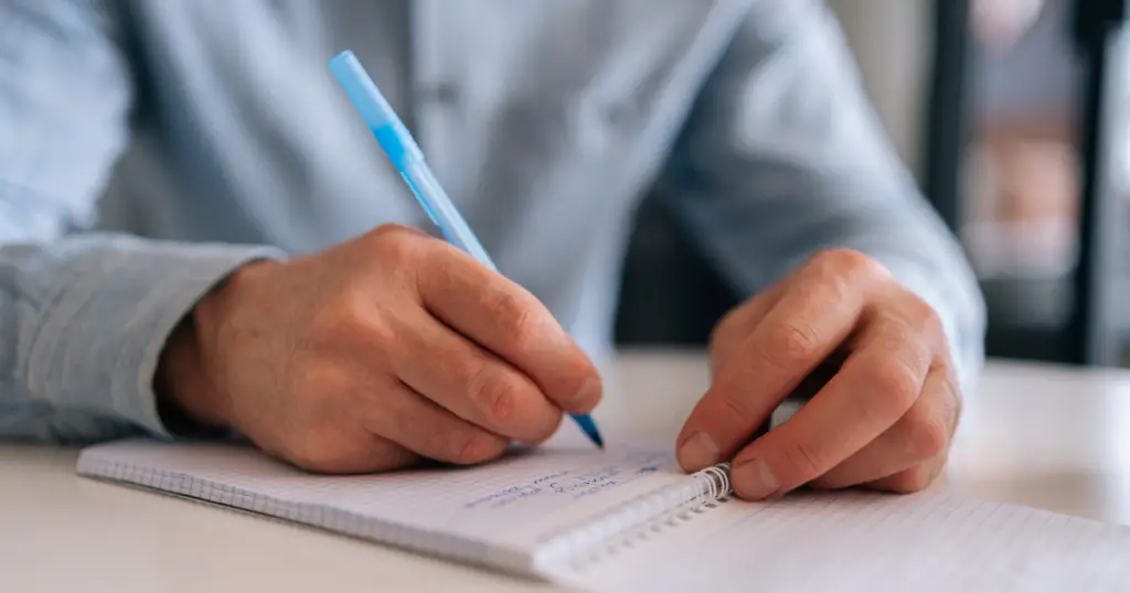 Person wearing a light blue button down shirt writing with a pen on a notepad